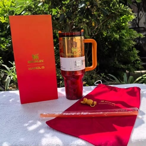 Red insulated tumbler with orange handle, red sleeve, and red box on a white surface with greenery in the background.