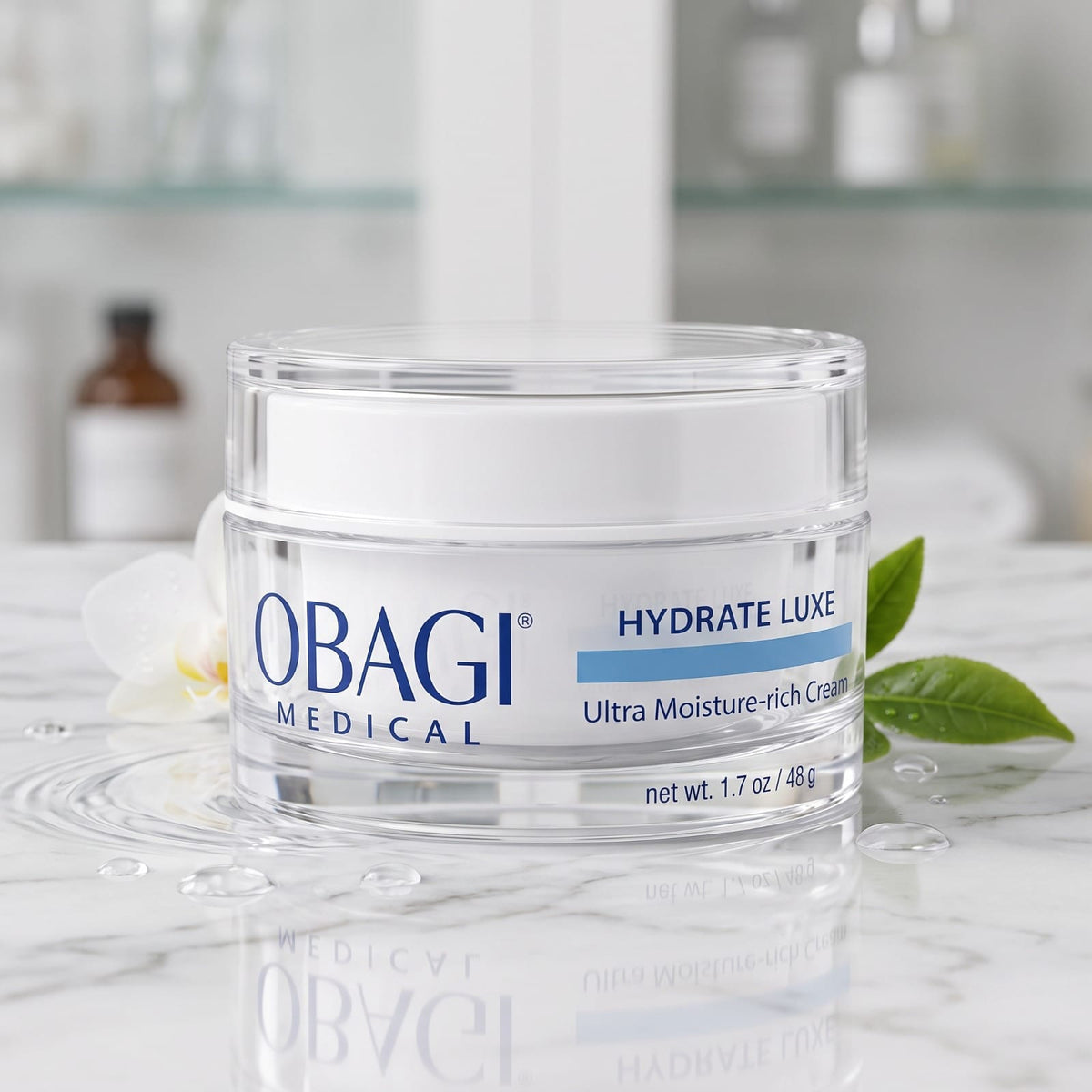 A clear cosmetic jar of Obagi Medical Hydrate Luxe Ultra Moisture-rich Cream on a white marble surface with a blurred bathroom-like background.