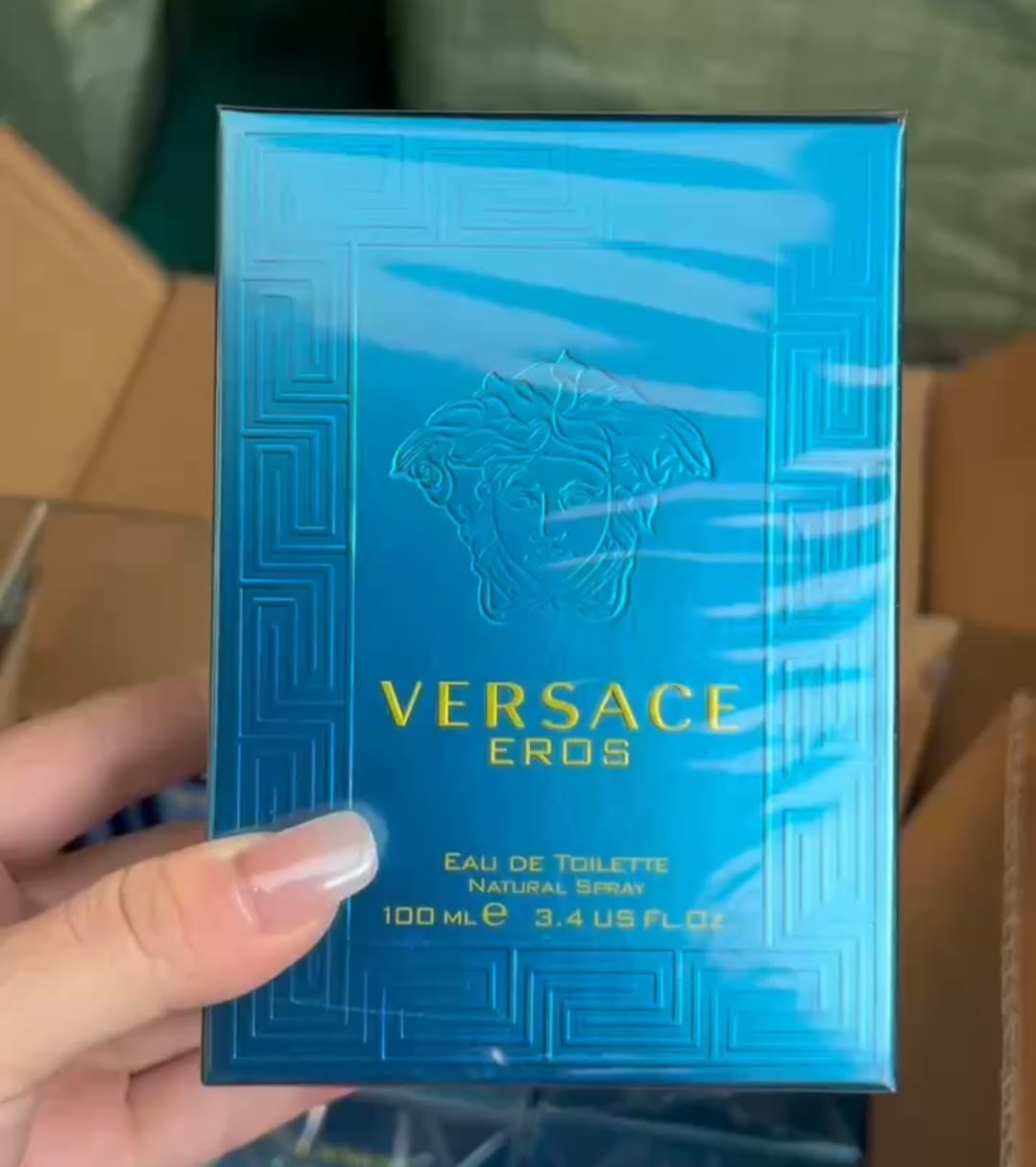 A hand holding a blue Versace Eros Eau de Toilette box wrapped in protective plastic, with cardboard boxes in the background.