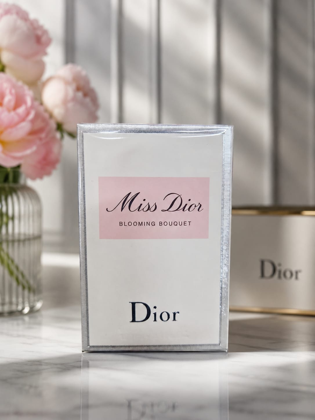 Dior Miss Dior Blooming Bouquet perfume box standing upright on a marble surface, with pink roses in a vase on the left and a blurred Dior container in the background.