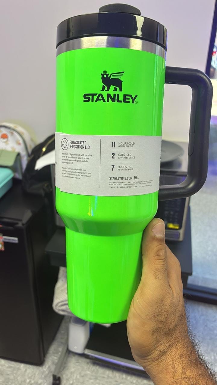 A bright green Stanley travel mug with a black lid and handle, held by a hand in an office setting. The mug features the black Stanley logo and a white product information label.