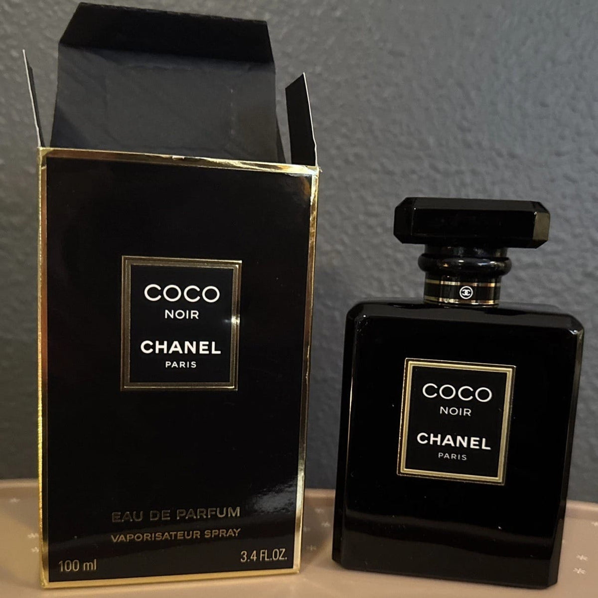 Chanel Coco Noir Eau de Parfum 100ml bottle and its original black and gold packaging box, displayed on a light surface against a gray wall.