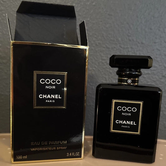 Chanel Coco Noir Eau de Parfum 100ml bottle and its original black and gold packaging box, displayed on a light surface against a gray wall.