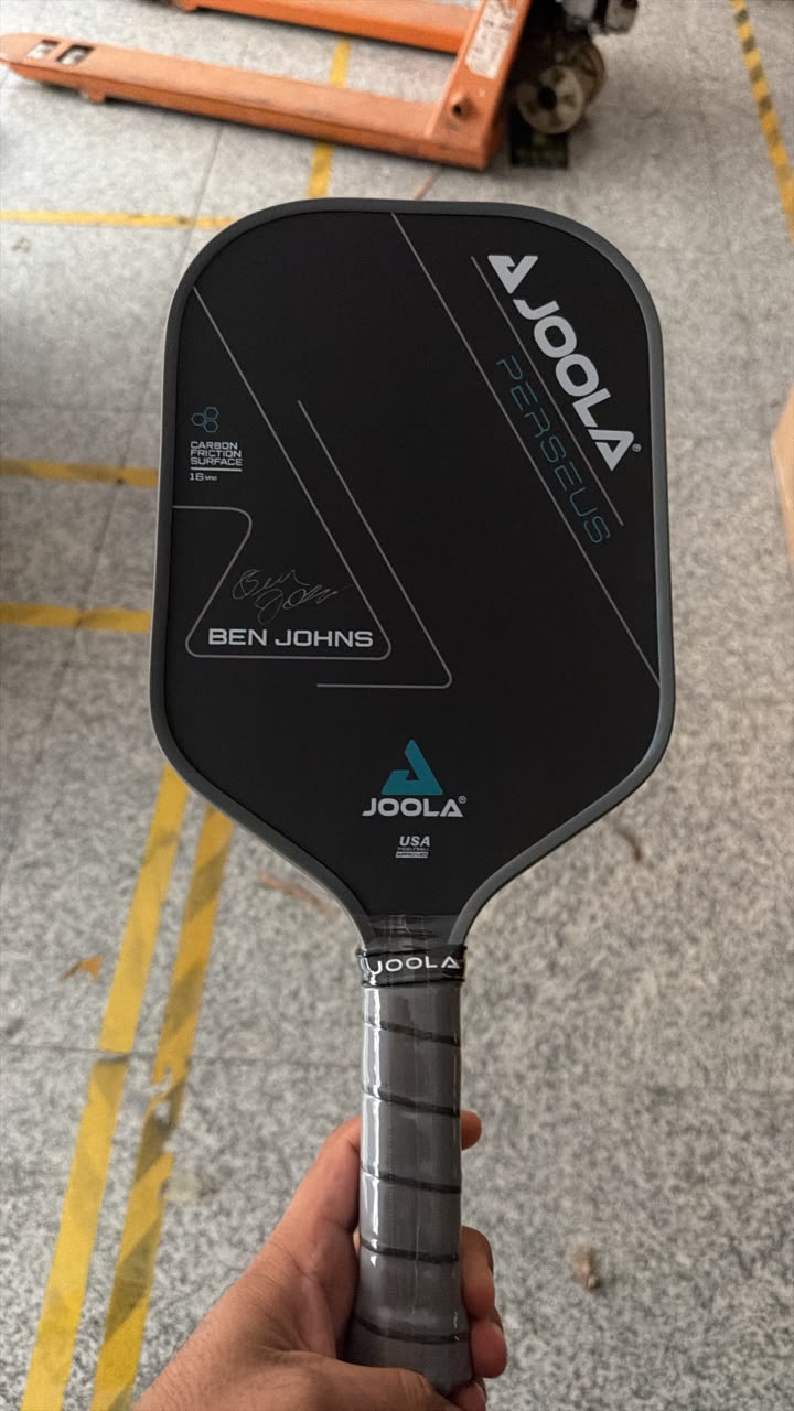 A hand holding a black Joola PENSUS Ben Johns pickleball paddle on a gray concrete floor with yellow painted lines.