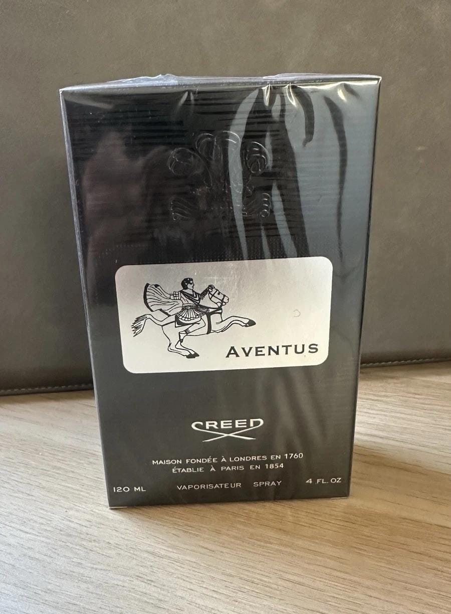 A boxed Creed Aventus fragrance package, photographed standing upright on a light wood surface against a dark gray background. The box is mostly black with a large white front label showing the Aventus horse-and-rider logo and the word AVENTUS, plus the Creed brand name near the bottom.