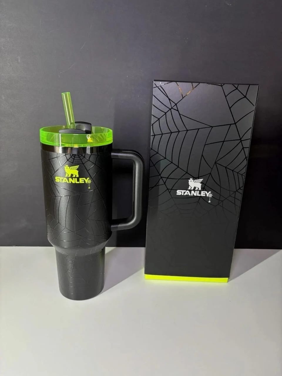 A black Stanley insulated tumbler with a handle, lid, and straw, shown next to its branded black box on a light surface against a dark background.