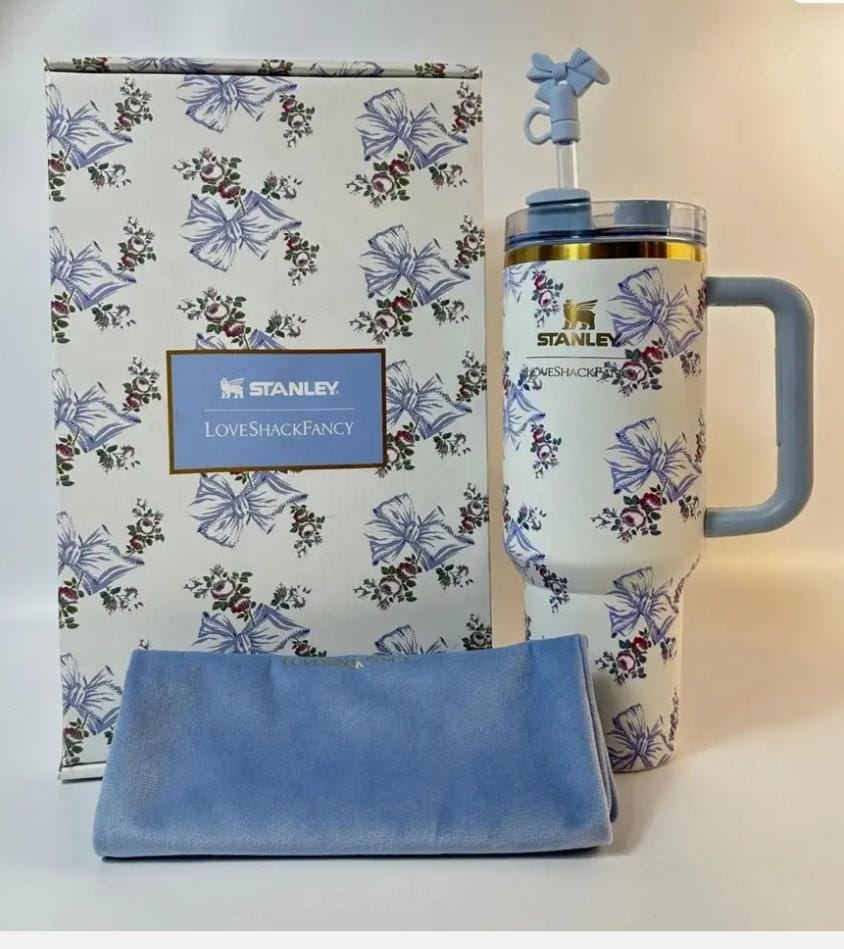 Stanley insulated travel mug with a blue bow and floral print, shown with its branded gift box and a blue microfiber cleaning cloth on a light beige background.